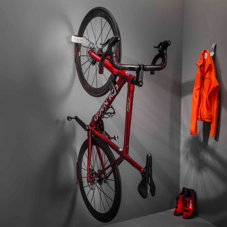 Colnago C64 road bike with red frame being stored vertically from the front wheel on a Cactus Tongue WRAP bike mount with a grey sleeve. An orange Le Col cycling is hanging on a Scoop Accessories hanger next to the bike. 